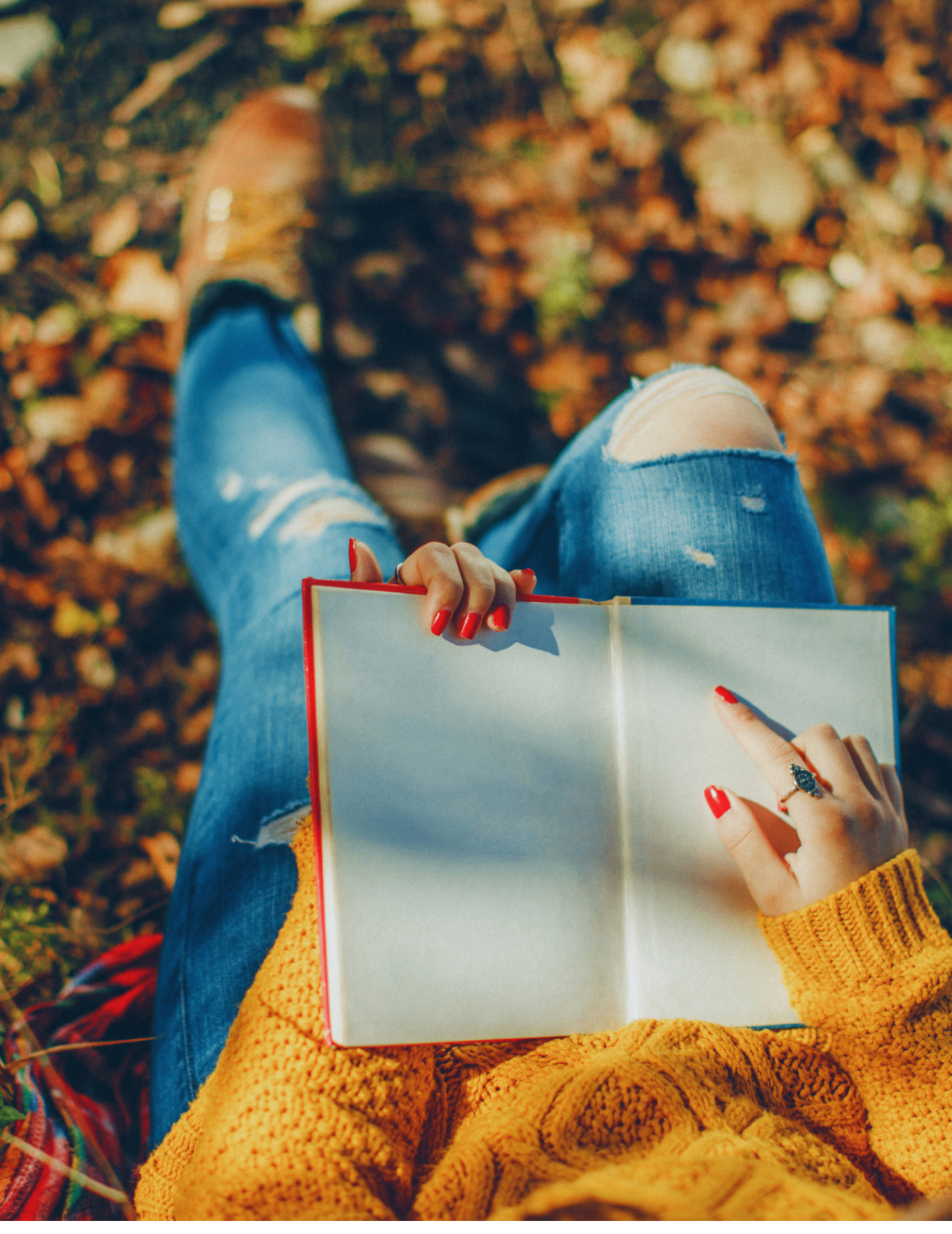 A woman in a mustard sweater reading an open book outdoors among autumn leaves